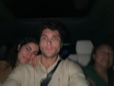 Three blurry people lie close in a dark interior. A man in a light button-down shirt has dark, curly hair and is partially obstructed by a black strap, possibly a seatbelt. To his right, a woman with curly hair smiles and touches her cheek. Another person is on the left, mostly out of focus and wearing a green shirt. They are likely in a car or similar vehicle.