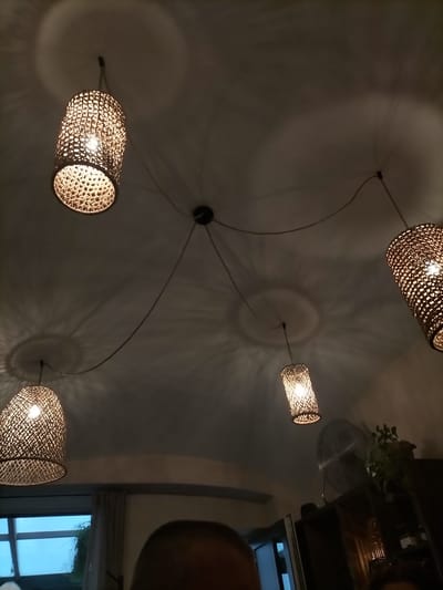 The image shows a dimly lit interior space with a ceiling from which four woven lamps are suspended by thin wires. The lamps cast intricate circular shadows on the ceiling. Two lamps are wider and shorter, while the other two are narrower and taller. The lighting is warm and creates a cozy atmosphere. In the lower portion of the image, a person's head is partially visible in the foreground, out of focus. To the right, a large fan and a shelf with plants and jars are visible. A window with a view of greenery is seen to the left. The overall scene suggests a restaurant or a domestic setting in Pardubice, Czechia.