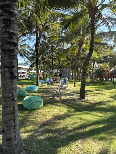 The image depicts a sunny outdoor scene at a resort in Cam Lâm, Vietnam. The foreground is dominated by the textured trunk of a palm tree on the left. Beyond, a lush green lawn is dotted with several large, mint green and teal beanbag chairs. In the mid-ground, a series of triangular white metal frames with colorful fabric hammocks hang from them – red, purple, and blue are visible. Behind these, a large, dark, rectangular screen or structure stands. Further back, rows of mature palm trees create a dense canopy, with glimpses of buildings and lounge chairs with white umbrellas visible through the fronds. The lighting suggests it is daytime, with strong sunlight casting distinct shadows of the palm trees across the grass.