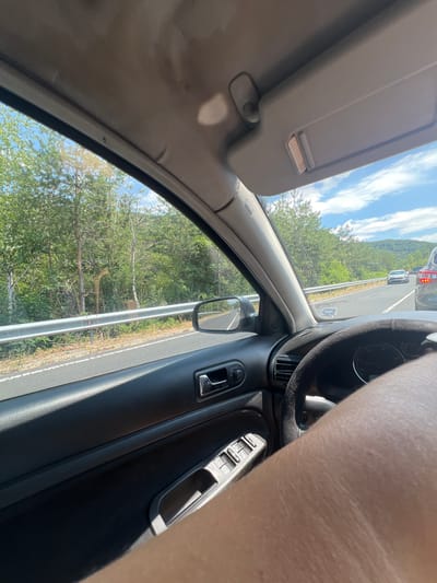 The image shows the interior of a car, looking out the driver's side window. The car is on a road with trees and a guardrail visible on the side. There are other cars on the road ahead, suggesting traffic. The sky is blue with some clouds. The interior of the car is visible, including the dashboard, steering wheel, and part of the driver's leg. The image appears to have been taken during the daytime. The location is described as Unknown, Bulgaria.