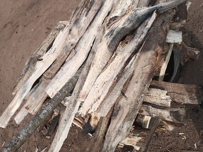 A pile of dry, weathered, and broken wooden branches are stacked on top of one another on dirt ground. The wood is grey, white, and black in color, and some of the branches are charred. The coordinates provided, 5.951 latitude and 5.697 longitude, are located in a rural area outside of the city of Adanisievbe, Nigeria. There are no people or other subjects in the image. The environment is a dry, dirt ground with no plants or other vegetation visible.