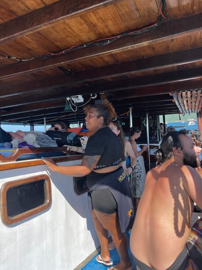 A group of people are on a boat. A person with curly brown hair is wearing a black shirt and shorts. They are leaning over the edge of the boat. Another person is lying down, shirtless, with their head towards the top of the frame. The boat has wooden walls and a white deck. Other people are relaxing or looking out at the water.