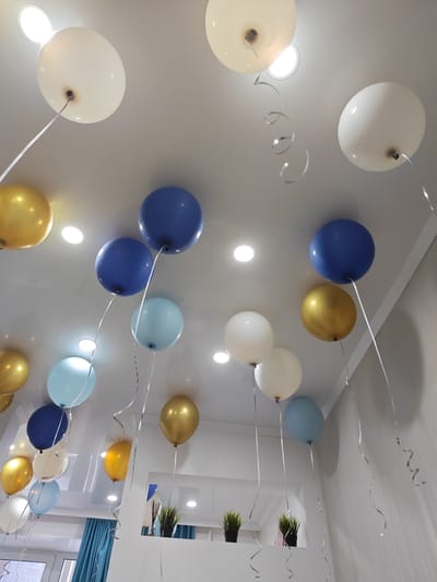 The image depicts a ceiling adorned with numerous balloons of various colors, including white, gold, blue, and light blue. Silver ribbons hang from the balloons, creating a festive atmosphere. Recessed lighting fixtures are visible on the ceiling, and in the lower portion of the image, glimpses of interior elements such as blue curtains, potted plants, and part of a wall can be seen. The scene appears to be an indoor celebration or party, possibly in a residential or event space in the city of Kumertau, Russia. There are no people or animals present in the image. The focus is entirely on the decorated ceiling and the balloons.