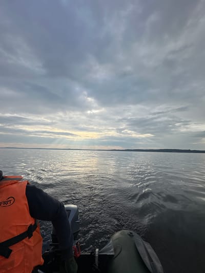 A low-angle, forward-facing shot from a boat on a lake shows a person wearing an orange life vest, their arm and shoulder visible as they operate an outboard motor. The boat moves through dark, choppy water, leaving a wake. In the distance, a tree-lined horizon separates the water from a cloudy sky. Shafts of light break through the clouds, illuminating the water. The scene is set on a lake near Shul'dikha, Russia.