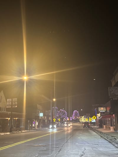 The image captures a nighttime street scene in what appears to be a town or city, heavily influenced by atmospheric conditions, likely fog or mist. The perspective is from the side of the road, looking down a street lined with buildings. The image is oriented vertically, with the road stretching into the distance on the right and the sky/fog on the left.
The dominant feature is the intense light diffusion and lens flares emanating from streetlights, indicative of fog or mist in the air. One very bright streetlight in the foreground creates a prominent starburst effect with strong horizontal and vertical rays of light. The street itself appears wet and reflective, with distinct double yellow lines marking the center. Patches of snow or slush are visible along the sides of the road and sidewalks.
Commercial buildings line both sides of the street. On the right side, signs are visible, including a brightly lit sign for "SEOUL KITCHEN" with a smiley face logo underneath, and smaller text reading "HOMEMADE AUTHENTIC KOREAN." Other less discernible business signs are also present.
Trees along the street are adorned with decorative lights; some are a vibrant purple, while others are yellow or white, suggesting a festive or holiday period. Several vehicles, appearing to be cars, are on the road, their headlights and taillights glowing through the misty air. Yellow diamond-shaped road signs with black arrows and pedestrian crossing symbols are visible along the roadside.
The overall scene conveys a quiet, atmospheric evening in a commercial district, with the fog enhancing the glow of the artificial lights. No people are visibly present in the frame. The time of day is clearly night. Given the provided context, the location is Leominster, United States.