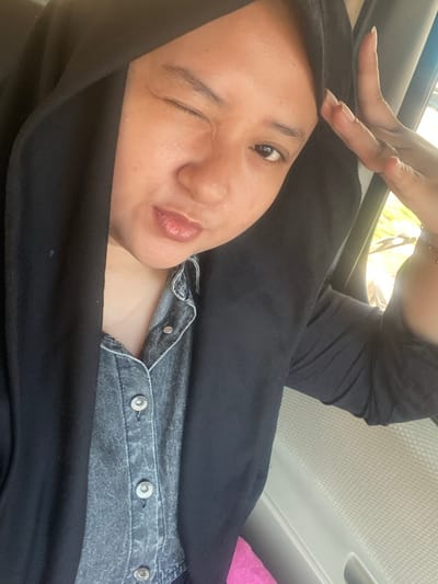 A young woman wearing a black hijab and a denim shirt is captured in a close-up, high-angle selfie. She is indoors, likely in a car, with the window visible on the right side. The woman is winking, with her right eye closed and her left eye open, and she has one hand raised near her forehead. The background suggests a blurred, natural setting with hints of greenery and sunlight. She is wearing a light-colored necklace and a bracelet on her right wrist. The image is focused on her face and upper body, highlighting her facial expression and attire. The location is identified as Ngrame, Indonesia.