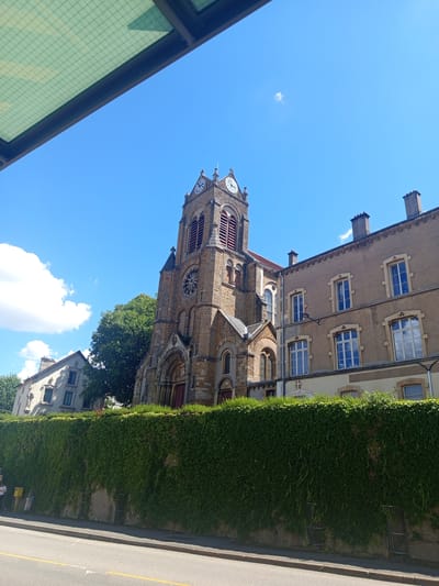 This media file depicts a street scene in Nancy, France, on a bright, clear day. The primary subject is a large, stone church tower with a clock face, visible from a low angle looking up into a vibrant blue sky. The tower features arched windows and intricate stonework. To the right of the tower, a tall, dense hedge of green ivy covers a wall, creating a verdant backdrop. Adjacent to the ivy-covered wall is a paved road with a yellow line, suggesting it's a public thoroughfare.
Behind and to the left of the church tower, another building with a lighter facade and multiple windows is visible. The windows on this building appear to be reflecting the blue sky. In the foreground on the left, a section of a structure with a green, grid-patterned glass pane is partially visible, possibly indicating a bus stop or a covered walkway.
The scene appears calm and tranquil, with no people actively engaged in any activity. The bright sunlight and clear blue sky suggest it is midday. The architectural style of the church and the presence of ivy are characteristic of many European cities. No visible text is discernible in the image. The overall impression is one of a peaceful urban setting on a sunny day, highlighting the historic architecture and lush greenery.
