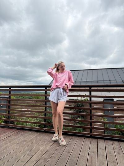 A young woman with blonde hair pulled into a ponytail stands on a wooden deck. She is wearing a pink long-sleeved sweatshirt and light grey shorts. Her right hand is in her hair. She is wearing light-colored clogs with pink and black designs. Behind her is a wooden railing and a dark roof of a building. Beyond the railing, there is greenery and a cloudy sky. The scene is outdoors, possibly on a balcony or deck overlooking a landscape. The location is identified as Semiluki, Russia.