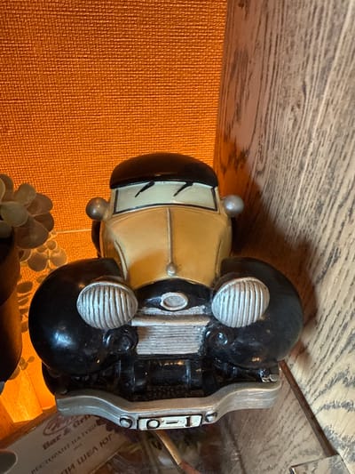 The image displays a close-up, top-down view of a decorative model car, oriented vertically with its front pointing upwards. The car, styled like a vintage automobile, has a light yellow or beige body, black fenders, and a black roof. Its most prominent features include large, circular silver headlights with horizontal grilles, a silver front grille with vertical slats, and smaller silver dome-shaped lights on the front fenders and possibly the roof. The windshield has black stylized markings, potentially representing wipers or a face. The rear bumper and surrounding structure are silver.
The model car rests on an unseen surface, with a textured, burnt orange material forming the background to its left and below. Above the car, a wooden surface with a distinct grain pattern is visible. Below the front of the car, out of focus, are parts of what appear to be artificial succulent plants or similar decorative foliage. To the right of the car, a blurry brown object or box shows partially visible text, largely in Cyrillic script. Phrases such as "Bar & Grill" and "РЕСТОРАНТ НА ТОЧНО" (Restaurant exactly/precisely) can be deciphered, along with other incomplete words like "ЖОИ ЯМ" (Joi Yam). The scene is static, depicting a collection of decorative items in what appears to be an indoor setting.