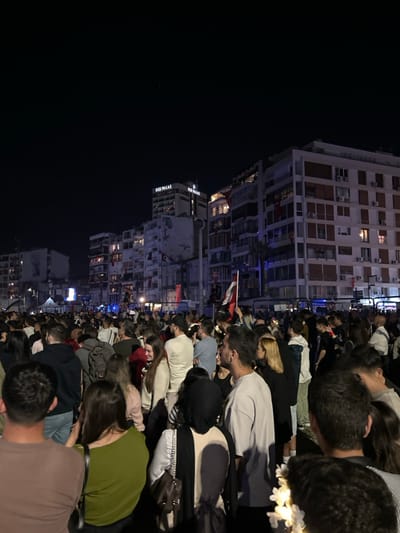 The image depicts a crowd of people gathered in front of a row of buildings in İzmir, Türkiye, at the coordinates 38.435, 27.139. The buildings are lit up by streetlights, with a large building labeled "İÇİ PALAS" prominent in the background. Several Turkish flags can be seen hanging from the buildings. The crowd consists of people of various ages and genders, dressed in everyday clothing. They are standing close together, and some are waving Turkish flags. The atmosphere is celebratory, with people smiling and talking to each other. The scene is well-lit and appears to be taking place at night. The crowd is facing a large building that is not visible in the image.