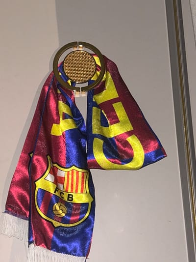 The image shows a FC Barcelona scarf displayed indoors. The scarf is predominantly red and blue with yellow detailing, featuring the FC Barcelona crest and the word "FCB". It is draped through a gold-colored circular object with a textured center. The background appears to be a plain, light-colored wall. The location, Topolovgrad, Bulgaria, is not visually evident in the image.