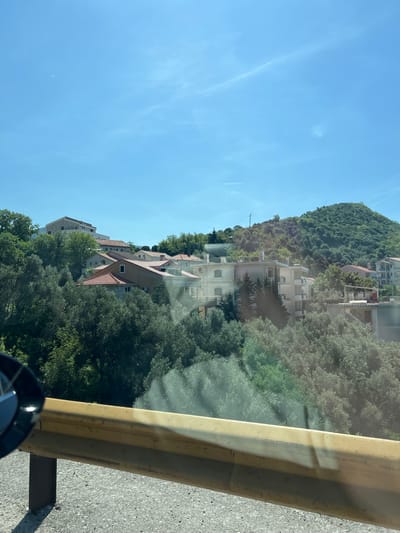 The image, taken from a moving vehicle, captures a bright, sunny daytime scene of a mountainous, verdant landscape with residential buildings, likely in Prijevorac, Montenegro.
In the immediate foreground, a yellow, weathered metal guardrail spans the right side, suggesting the vantage point is from a road or elevated viewpoint. A reflective surface, likely a car window, shows subtle distortions and reflections across the scene, particularly in the sky and on the guardrail itself. Below the guardrail, a dense canopy of green trees and shrubbery fills the lower and mid-section of the frame, hinting at a valley or steep hillside.
Amidst the lush greenery and rising up the slopes are several multi-story residential buildings. These structures feature light-colored facades, predominantly white or cream, with distinct reddish-brown tiled roofs. Many of them include balconies and multiple windows, suggesting apartment complexes or larger homes nestled into the hillside.
Dominating the upper left and central background is a large, imposing hill or mountain, entirely covered in a thick, dark green forest of trees. The sky above is a clear, vibrant blue, with a few faint, wispy contrails visible, indicating a serene and cloudless day. The overall impression is one of a picturesque, sunny day in a mountainous coastal or hilly region, with development integrated into the natural landscape. No people or visible text are present in the image.