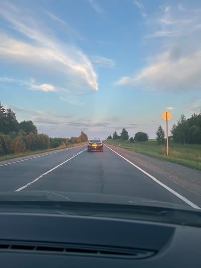 The image captures a view from inside a vehicle looking out at a road in Glazunovo, Russia. The scene depicts a two-lane asphalt road with white markings, bordered by grassy shoulders and a dense line of trees on the left and right. In the distance, a dark-colored car with its license plate illuminated is driving ahead. The sky above is a blend of blue and white clouds, with sunlight casting a warm glow. A circular yellow sign is visible on a pole on the right side of the road. The foreground shows the dashboard of the vehicle from which the photo was taken.