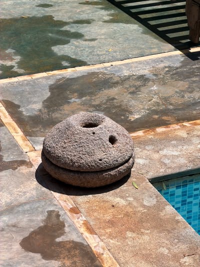 The image displays an outdoor scene in Tahannaout, Morocco, featuring a large, roughly carved stone object resting on a textured concrete or stone patio. The stone object appears to be composed of two stacked, disc-shaped sections, resembling a large millstone or a decorative garden element. Its visible surface displays a deep, roughly circular hole near the center and two smaller, shallow holes towards the top.
Adjacent to the stone object, the clear blue square tiles of a swimming pool are visible, indicating a poolside or courtyard setting. The surrounding concrete or stone floor exhibits signs of wear, with varying textures, cracks, and patches of dampness or discoloration.
Strong, well-defined shadows, including a pattern of parallel bars from an unseen structure (likely a fence or railing) in the upper left, suggest bright sunlight, possibly during midday or early afternoon. The overall atmosphere is still and calm, with no human activity or visible text present.