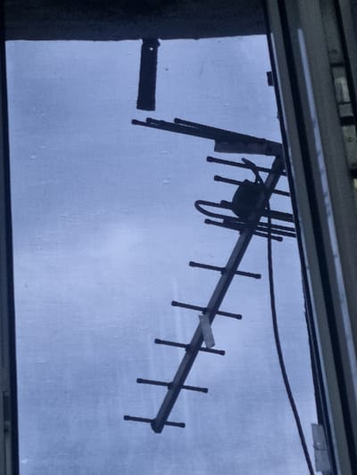 The image shows a close-up, angled view of a television antenna, likely a Yagi-Uda antenna, positioned against a pale blue, overcast sky. The antenna is dark in color and appears to be made of metal. It is mounted on a vertical pole or mast, which is partially visible. The antenna has multiple horizontal elements of varying lengths extending from a central boom. A small, dark box-like component is attached to the boom. A dark cable runs from the antenna down and out of frame. The scene is framed by what appears to be a window or door frame on the right side of the image, which is a lighter, neutral color and has vertical lines, suggesting a building structure. The sky has a slightly textured or mottled appearance, possibly due to rain or condensation on a windowpane. There are no people or other discernable objects in the image. The overall impression is one of an outdoor or semi-outdoor setting, common in residential or urban environments where television reception is enhanced by such antennas. The location is specified as Shopokov, Kyrgyzstan.