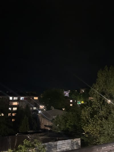 The image depicts a nighttime scene in Gladzor, Armenia. The view is from a slightly elevated position, looking down and to the right at a residential building and surrounding trees. The sky is completely dark, indicating it is nighttime. The building, partially obscured by trees and shadows, has several windows illuminated with warm yellow light, suggesting that residents are inside. The trees are dense and leafy, with some branches extending into the frame and casting shadows. The overall atmosphere is quiet and peaceful, typical of a residential area at night. There is no visible text in the image.