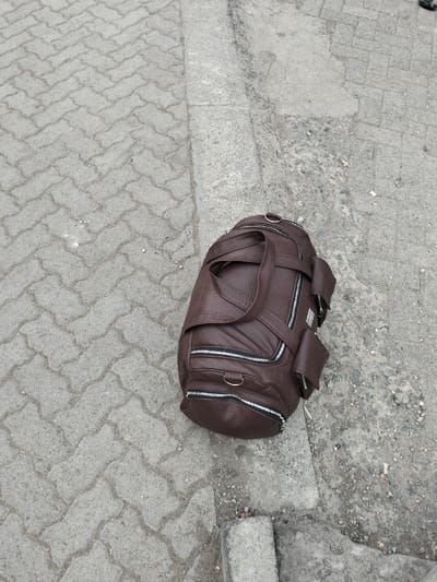 A brown leather duffel bag rests on a grey curb and adjacent cobblestone sidewalk. The bag features several zippered compartments and carrying handles. The curb separates the cobblestone from a concrete surface with scattered debris. The bag is positioned at an angle, taking up a significant portion of the right side of the frame.