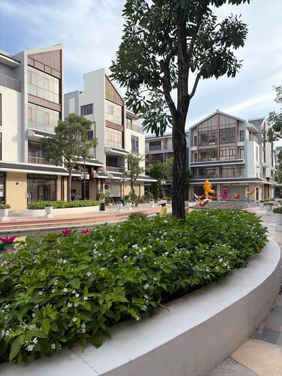 The image depicts a modern urban development in Nghĩa Trụ Commune, Vietnam, featuring a row of multi-story buildings with distinctive architectural elements. The buildings are white with grey accents and feature large windows with wooden frames, and some have angled, slatted wooden panels on their upper facades. In front of the buildings is a wide, paved walkway alongside a narrow canal or water feature.
A lush, manicured hedge of green bushes with small white flowers lines the edge of the walkway overlooking the water. The water feature contains several colorful, geometric origami sculptures, including pink flowers, yellow birds, and a large yellow cartoon character resembling Winnie the Pooh, as well as a pink sculptural element.
A large tree with a dark trunk and dense green foliage is prominently positioned in the foreground, partially obscuring the view of the buildings. The ground is paved with interlocking bricks in shades of red, grey, and yellow. A few white potted plants are visible along the walkway. In the middle ground, a person wearing a conical hat and a yellow outfit stands near the water feature. The sky is overcast with a few wispy clouds. The overall scene suggests a commercial or residential complex with a focus on aesthetic landscaping and public art.