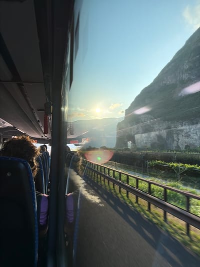The image shows a view from inside a bus traveling on a road in Roveré della Luna, Italy. The sun is setting, casting a warm glow over the mountainous landscape. A steep, green-covered mountain dominates the right side of the frame, with a rocky cliff face visible. In the foreground, a metal guardrail runs alongside the road, bordered by lush green vegetation, likely vineyards. The road surface appears to be asphalt, showing motion blur indicative of the bus's movement. Inside the bus, passengers are visible, with the back of their heads and shoulders showing. One passenger in the foreground has curly brown hair. The seats are upholstered in blue fabric with a patterned design. The bus interior features overhead compartments and lighting fixtures. The scene is bathed in the golden light of the setting sun, creating long shadows on the road.