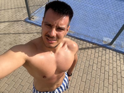 A man is taking a selfie outdoors. He is shirtless and wearing blue and white striped swim shorts. He is standing on a paved walkway with a blue surface and a chain-link fence behind him. The lighting suggests it is daytime. The location is Roquetas de Mar, Spain.