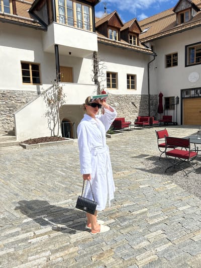 A woman stands in the courtyard of the Hotel Restaurant Schwarzer Adler in Pflaurenz-Floronzo, Italy. She wears a white bathrobe, white shoes, and sunglasses, and carries a black Chanel purse. She holds a book over her head. The courtyard is paved with gray stones, and red patio chairs and tables stand nearby. Buildings of white stucco and wood frame the courtyard. The sky is blue with a few clouds.