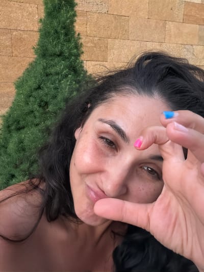 The media file features a close-up, rotated shot of a person's face as they form a heart shape with their hands over their eye. The person has dark hair and is looking towards the camera with a gentle expression. Their fingernails are painted in two different colors: blue on one finger and pink on another. The background shows a textured surface of tan tiles and a section of dense green foliage, possibly grass or a bush. The lighting suggests it might be daytime, possibly outdoors. The image has a warm tone and a slightly soft focus, giving it a personal and intimate feel. There is no visible text in the media file.
