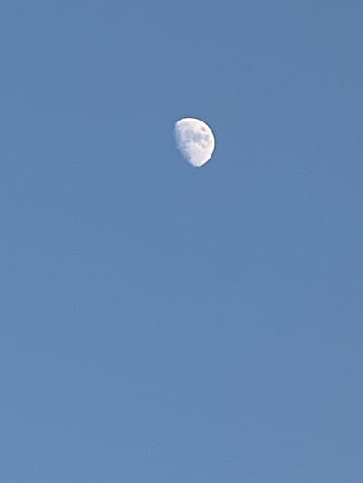 The image depicts a daytime view of the sky with a gibbous moon visible. The sky is a clear, uniform blue, suggesting fair weather. The moon is partially illuminated, with its shadowed portion on the right side. Surface details of the moon, such as craters and mare, are discernible. There are no people or man-made objects visible in the frame, focusing solely on the celestial body against the backdrop of the sky. The lighting suggests it is daytime, as the sky is blue and the moon is visible. The image appears to be a photograph taken from an outdoor location.