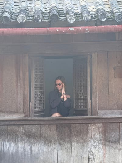A woman wearing sunglasses and a dark long-sleeved shirt looks out of a window with a lattice grill. The structure of the window and surrounding walls is wooden. Old, patterned, ceramic roof tiles line the edge of the frame to the left. The scene is in Sà Phìn Commune, Vietnam.