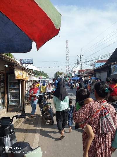 A bustling street scene unfolds under a partly cloudy sky. A tall communication tower rises in the distance. Various individuals traverse the street, some on foot and others on motorbikes. A woman carries a child on her back. Shops and stalls line the street, shaded by awnings. An umbrella with red, white and blue colours hangs over the scene. A scooter is parked on the side of the road.