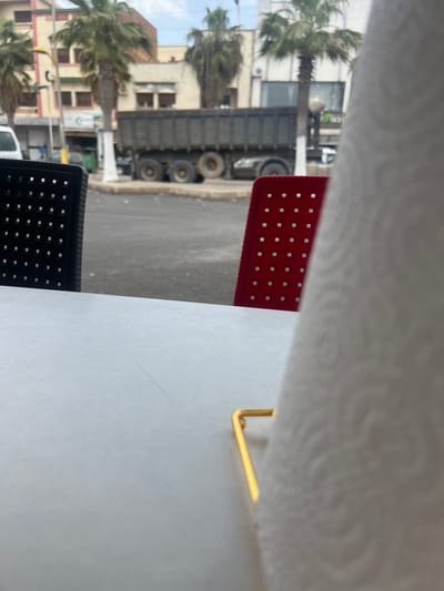 The image presents an outdoor scene, likely viewed from a patio or balcony in Boufakrane, Morocco, looking out onto a street and buildings. In the immediate foreground, a white tabletop or surface is visible, partially obscured by what appears to be a textured white paper towel roll held by a golden metal bracket on the right. To the left of this, parts of two outdoor chairs are seen: a red chair with a pattern of small square perforations, positioned higher in the frame, and a black chair with a similar perforated design, located lower.

The midground features a street with various vehicles. A prominent, large dark grey truck, possibly a dump truck or similar heavy vehicle, is parked or driving on the street, its wheels clearly visible. Other vehicles, including a white van, are also discernible. Along the street and in the background, several multi-story buildings with light-colored, possibly beige or brown, facades and numerous windows line the area. Some buildings feature external air conditioning units. Scattered throughout the scene are numerous palm trees, their green fronds adding a tropical touch to the urban landscape.

The sky is bright, suggesting daytime, likely overcast or partly cloudy, as there are no harsh shadows. A green sign with white text, which appears to be Arabic script, is vaguely visible on one of the buildings, though the text itself is too blurry to read. The overall impression is a typical daytime street scene in a North African town.