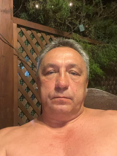The image shows a close-up selfie of a middle-aged man, likely taken outdoors in the evening. The man is shirtless and his upper body is visible, with his face angled towards the camera. He has graying hair and appears to be in good health.
Behind the man, a wooden lattice fence is visible, with greenery behind it. The lighting suggests it is nighttime, possibly illuminated by artificial lights, as evidenced by some faint, out-of-focus lights in the background foliage. The overall setting appears to be a garden or patio area. The man's expression is neutral, and there are no overt signs of specific activity or emotional distress. There is no visible text in the image. The location context places this scene within the Germasogeia Municipality in Cyprus.