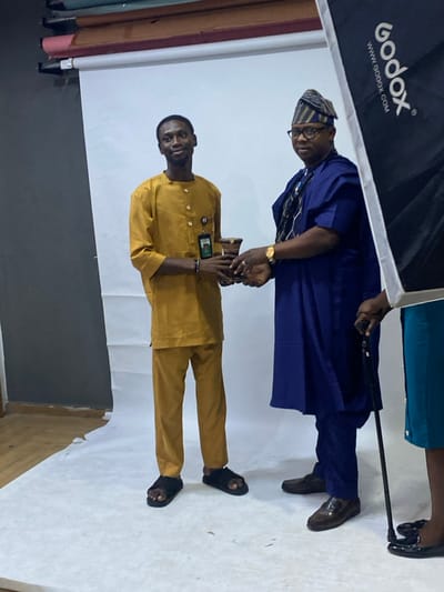 Two men stand together in a studio setting. The first man, dressed in a mustard-colored traditional outfit and sandals, stands in front. The second man wears a navy blue outfit with a patterned cap and dark shoes, holding a black stick. The backdrop is a white studio setup with Godox lighting equipment visible. Their hands are clasped.