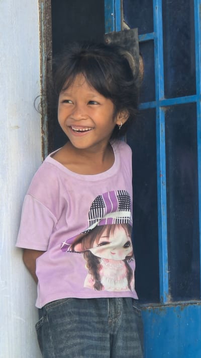 A smiling young girl with dark hair and an earring is lying on her side, wearing a light purple t-shirt with a graphic print, and denim pants. She is positioned against a blue metal grate. The setting suggests an outdoor urban location, consistent with Ba Ngoi Ward, Vietnam.