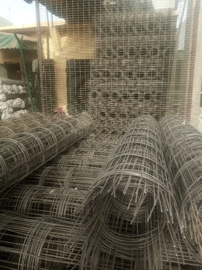 The image depicts a construction material storage area in Kofar Ruwa, Nigeria. The foreground and midground are dominated by numerous rolls of dark gray metal wire mesh, likely welded or woven steel, stacked horizontally on what appears to be ground level. These rolls vary in diameter and length, some showing exposed wire ends. In the background, a large quantity of similar wire mesh is stacked vertically against a light-colored building wall, forming a grid-like pattern across much of the upper portion of the frame. To the left, additional bundles of material, possibly similar mesh wrapped in white protective covering, are visible. Part of a building structure with a green awning is present on the far left, suggesting an outdoor or semi-enclosed commercial setting. No individuals or active interactions are visible in the scene. The lighting indicates daylight conditions.