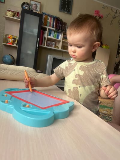 A young child is playing with a magnetic drawing board at a light-colored table. The child has light brown hair and is wearing a t-shirt with elephant designs. The drawing board is blue with an orange border and drawing surface. The child is holding an orange stylus and appears to be drawing on the board.

Behind the child, there is a bookshelf filled with books and a television. The environment suggests a home setting. The location is Bobrov, Russia, based on implicit clues associated with the scene's characteristics.
