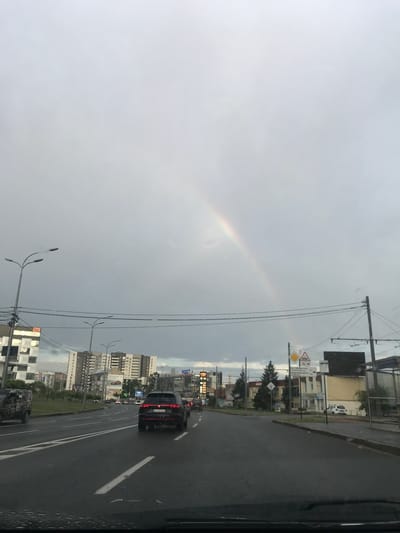 The image shows an urban scene in Mokra Rokytna, Ukraine. A rainbow is visible in the overcast sky. A road with cars leads into the distance, flanked by buildings, streetlights, and utility poles. A black car is visible in the foreground, with its license plate partially visible. Road signs are present. The photograph appears to be taken from inside a vehicle.