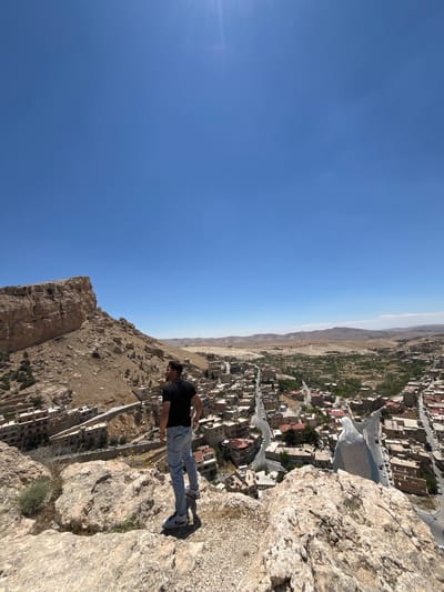 A man stands on a rocky cliff overlooking Ma'loula, Syria. He wears a dark t-shirt, blue jeans, and dark shoes. The city is visible in the background. The sky is clear and blue.