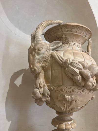 The image shows a detailed, close-up view of an ornate, light-colored vase or urn. The vase features a sculpted ram's head with prominent horns and beard, positioned on its side and appearing to emerge from the body of the vessel. Draped around the vase are garlands of fruit or seed pods. The vase itself has a ribbed body and is decorated with intricate patterns around its base and upper rim, including acanthus leaves and geometric motifs. The object is lit from the side, casting a strong shadow on the textured, off-white wall behind it. The vase appears to be made of stone or a similar material and exhibits some signs of age or wear, with visible cracks on the rim and surface. The lighting and setting suggest an indoor environment, possibly a museum or a historical building in Évian-les-Bains, France.