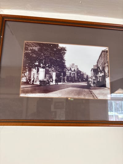 This framed, sepia-toned historical photograph depicts a street scene identified by its caption as Church Street, Oswestry, United Kingdom, likely from the early to mid-20th century. The wide street recedes into the background, flanked by buildings and trees.

On the left side of the street, a large, mature tree with dense foliage partially obscures elaborate stone gateposts and ornate iron gates. Approximately five to seven individuals, dressed in period attire, are visible near these gates and beneath the tree. A vintage open-top automobile, possibly a tourer with visible side-mounted spare tires, is parked facing the right near the left curb, with at least one occupant in the driver's seat. Beyond the tree, a multi-story building featuring distinct half-timbered construction is visible amongst other structures.

The right side of the street is lined with a continuous row of multi-story brick buildings, appearing to be commercial or residential properties, some with ground-level shopfronts. Several pedestrians are positioned along the sidewalk on this side. Further down the street on the right, another indistinct vintage vehicle is partially visible.

The paved roadway occupies the central foreground and midground, leading into the distance. A tall street lamp stands on the left side of the street. Approximately 10-15 individuals are dispersed throughout the scene, either standing or walking on the sidewalks.