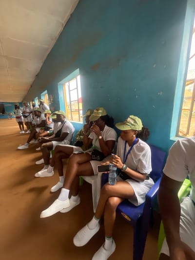 In Ise-Ekiti, Nigeria, a group of young people are seated in a row, facing right, within a room that appears to be a communal area or classroom. The walls are painted a vibrant teal blue, with a stark white ceiling visible above. Sunlight streams in through a large window on the right side of the image, illuminating the scene and casting shadows. The floor is a warm, reddish-brown earthen tone.
The individuals are dressed uniformly in white t-shirts, white shorts, and light green caps. Many of them are wearing white socks and white sneakers. Some are holding personal items, such as mobile phones and water bottles.
The group appears to be composed of both males and females. On the far left, a woman stands, and to her right, several individuals are seated on chairs that are primarily blue with some green accents. The seating arrangement suggests a waiting or gathering situation. The individuals are generally looking forward or downwards, with some interacting with their phones. One woman in the foreground is holding a water bottle. The overall atmosphere seems calm and orderly.