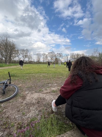 A medium-shot, tilted outdoor image. A person wearing a black puffy vest, a maroon hoodie, black leggings, and white sneakers is partially visible on the right. Their curly brown hair is also visible. In the distance, a group of people are standing on a green field. Bare trees are scattered throughout the field. A bicycle is in the lower right foreground. The sky is visible in the upper left, with a mix of blue sky and white clouds. The location appears to be a park or grassy area. There is no information in the image to suggest that the location is Gromshin, Bulgaria.