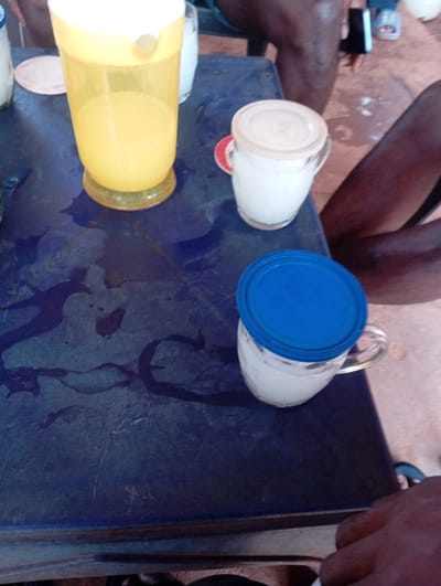 The scene depicts a dark blue, possibly plastic, table surface in Okpatu-Ukehe, Nigeria, observed from a high-angle perspective. On the table, a tall, transparent yellow plastic pitcher with a white lid contains a yellow liquid. To its right, a clear glass mug with a beige plastic lid holds a white opaque liquid and rests on a circular red and white coaster. Further towards the bottom right, another clear glass mug with a bright blue plastic lid also contains a white opaque liquid. The table surface exhibits multiple dark liquid spills and scratches. Partially visible in the upper right background are the dark-skinned legs of at least one individual, one leg bent and wearing a dark garment, with a foot in a light-colored sandal visible further back. In the lower right foreground, a dark-skinned arm and hand are visible, suggesting another individual is seated at the table, with a foot in dark flip-flops or sandals also partially discernible.