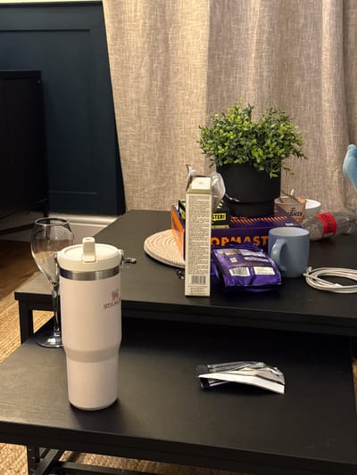 The image shows a cluttered black table indoors. On the table are various objects including a small potted plant, a blue mug, a box of Topmaster tools, several chocolate wrappers, a bottle of Coca-Cola, a white insulated Stanley cup, and a wine glass. The background includes a beige curtain, a dark blue wall with paneling, and a television. The setting and objects do not provide enough information to confirm the location is Windermere, United Kingdom, or provide further details about people, actions, or interactions.