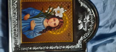 The image shows a framed icon of a young woman with brown hair wearing a blue robe and holding white lilies. The icon is set against a textured gold background with a circular, ornate yellow border. The frame is ornate and silver-toned, with detailed decorative elements. To the right of the icon, a portion of another decorative silver object is visible. The entire object rests on a blue, textured fabric. The image does not provide any information about the city of Bocani, Moldova.