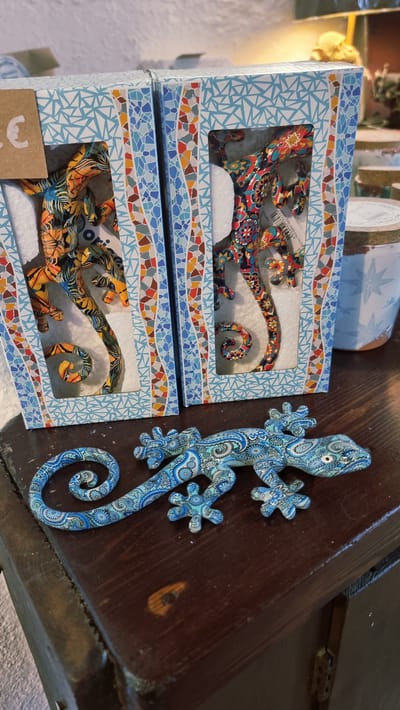 The image shows two decorative gecko figurines inside their boxes, positioned on a wooden surface. In the foreground, a third, larger gecko figurine with a blue paisley pattern is displayed on the wooden surface. The boxes have a geometric blue and white pattern with colorful mosaic-like borders. The gecko inside the left box is predominantly orange and yellow, while the one in the right box displays a multi-colored pattern. The background is out of focus but suggests an indoor setting, possibly a shop. The presence of a price tag with a euro symbol on the left box indicates a commercial environment. The scene appears to be a display of decorative items.