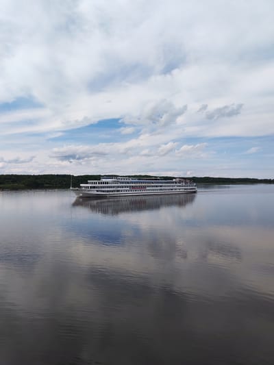 A wide, long, white, multi-deck cruise ship travels on a calm river towards the right side of the frame. The ship has numerous windows along its sides and a white superstructure. The water reflects the ship, the sky, and a distant tree line. The sky is filled with scattered white and gray clouds. The scene appears to be set in Shchetinino, Russia, on a river.