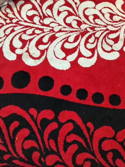 This is a close-up, overhead shot of a rug with a red background and a pattern. The pattern is divided into three horizontal sections. The top section features a swirling white design on red. The middle section is solid red with a row of black dots. The bottom section has a black background with a red swirling pattern. The rug appears to be indoors, possibly in a home. There are no people or other significant objects visible in the image. The location in the city of Cottrueng, Indonesia is not discernable from the image itself.