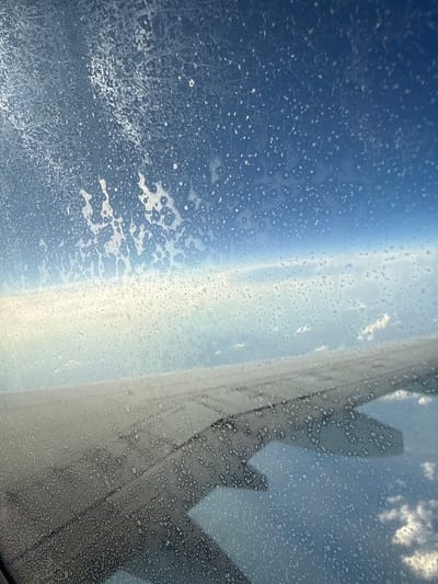 The image shows a view from an airplane window, looking out at the sky and clouds. The window is covered in condensation or frost, which creates a pattern of water droplets and ice crystals. The wing of the airplane is visible in the foreground, extending across the bottom of the frame. The sky is a deep blue, with white clouds scattered throughout. The overall impression is one of being high up in the atmosphere, looking out at a vast and cloudy expanse. There is no indication of the city of Vykhodnoi, Russia in this image.