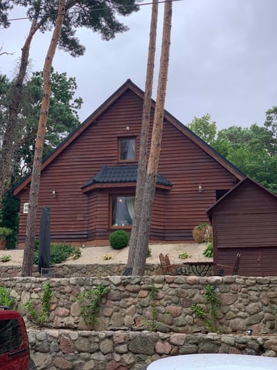 The image shows a wooden house in the city of Pobierowo, Poland. The house has a dark brown, horizontal wooden siding and a dark, tiled roof. There are two windows visible on the front of the house, one smaller upper window and a larger lower window. A dark-colored outdoor umbrella is leaning against the side of the house. In front of the house, there is a low, multi-layered stone wall. Behind the wall, there is a small outdoor seating area with a table and two chairs. There are several tall pine trees in the foreground and background, as well as lush green foliage. The sky is overcast. A red vehicle is partially visible in the lower left corner of the image.