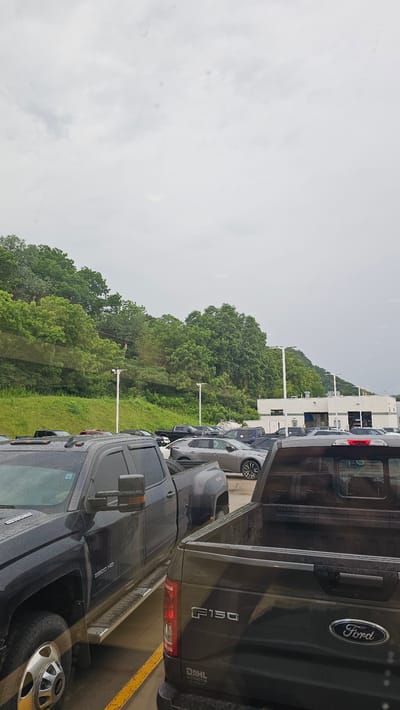 The image shows a parking lot filled with vehicles under a cloudy sky. In the foreground, two dark-colored pickup trucks are prominently visible. The truck on the left is a Chevrolet Silverado HD, and the truck on the right is a Ford F-150. Behind these trucks, there are rows of other cars and trucks, many of them appearing to be for sale, suggesting a car dealership. In the background, a lush green hillside covered with trees rises behind the parking lot, and a building with large windows and garage doors, likely part of the dealership, is visible to the right. The scene appears to be in Winona, Minnesota, United States, based on the context provided. The ground is wet, indicating recent rain.
